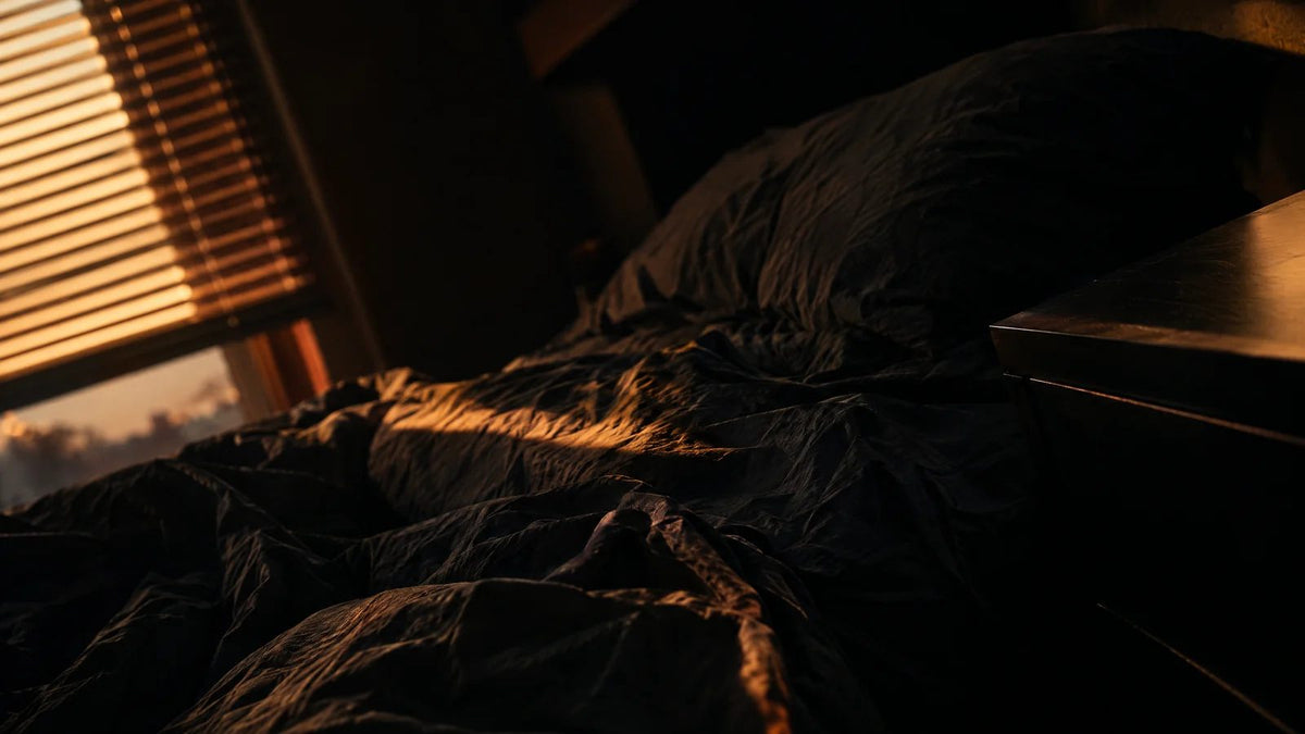 Rumpled bed in a dim bedroom with early morning light coming through the blinds